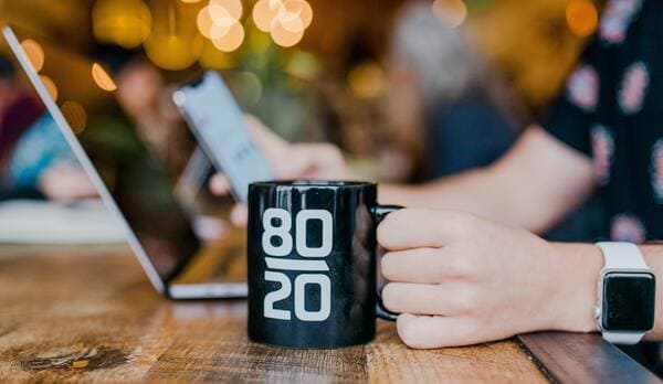 A picture containing text, coffee mug, person, hand, table, laptop, smartwatch, and a mobile phone.